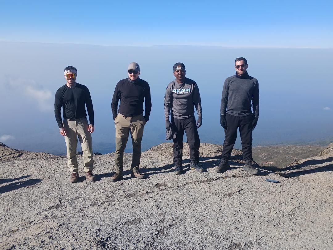 Joseph, third from left in a grey top and backwards cap, with three hikers in base layers on a sunlit rock ledge above a flat sea of cloud