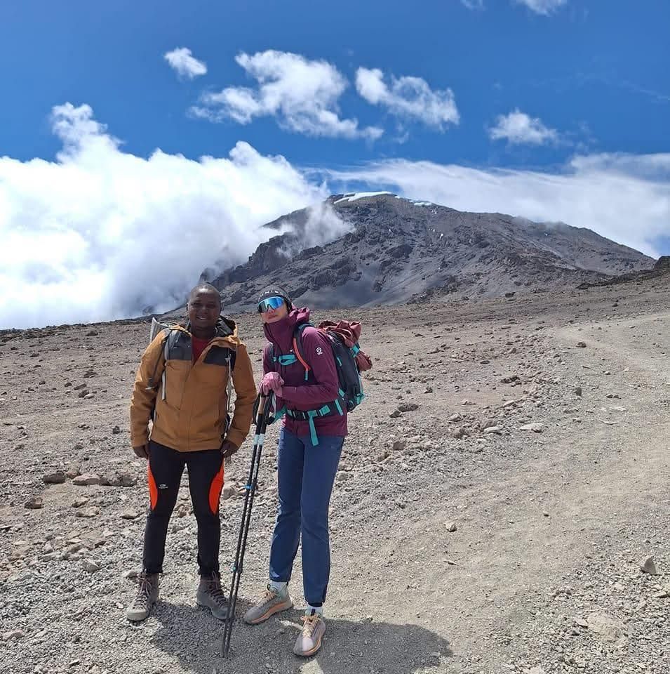 A guide and a hiker with trekking poles stand on a barren gravel plain below the rocky, cloud-wrapped summit