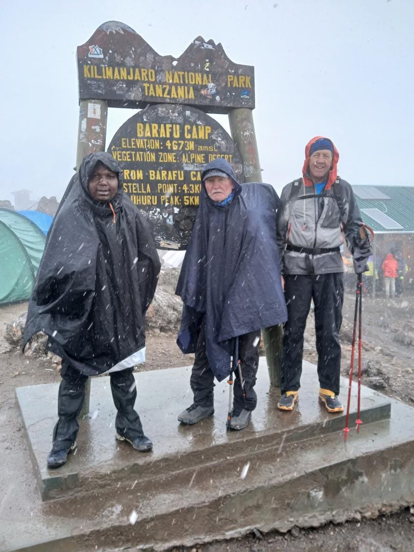Joseph, on the left in a black rain poncho, with two climbers in rain ponchos at the Barafu Camp sign, elevation 4673 m, in falling snow