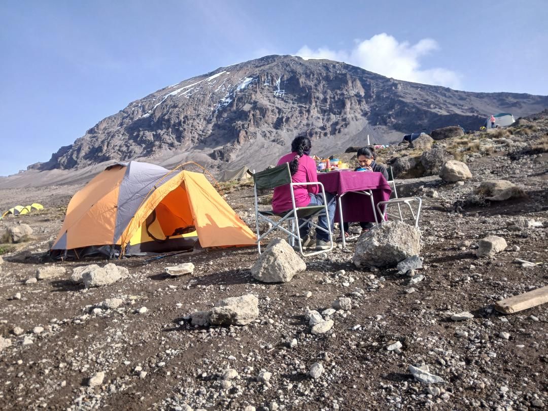 Two hikers eat at a camp table beside an orange tent on gravel ground, below a rocky, snow-streaked summit