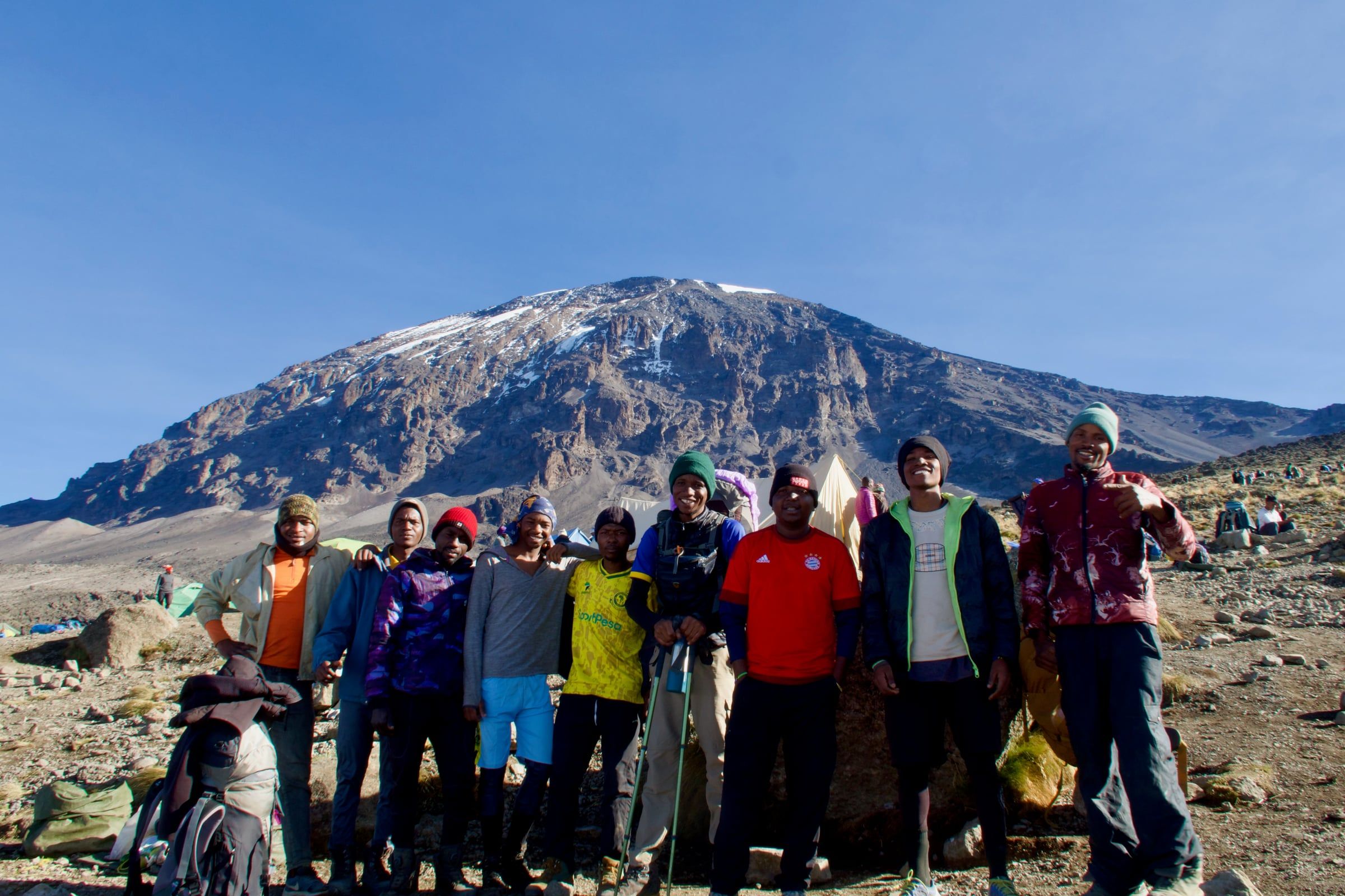 Mountain crew members smiling arm in arm on rocky ground below Kilimanjaro's snow-streaked summit cone. Joseph is in the red football shirt