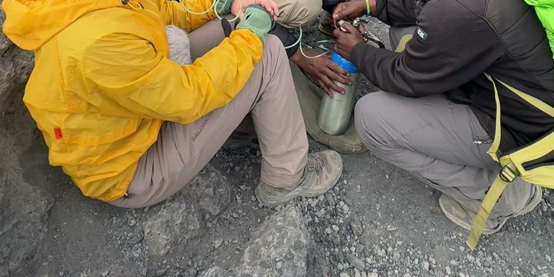 Crew members adjust a bottled-oxygen regulator for a seated climber in a yellow jacket on rocky ground