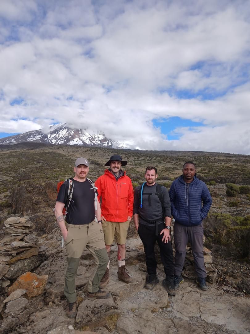 Four hikers on a rocky path across low scrubland, with a snow-capped peak half hidden in cloud behind them